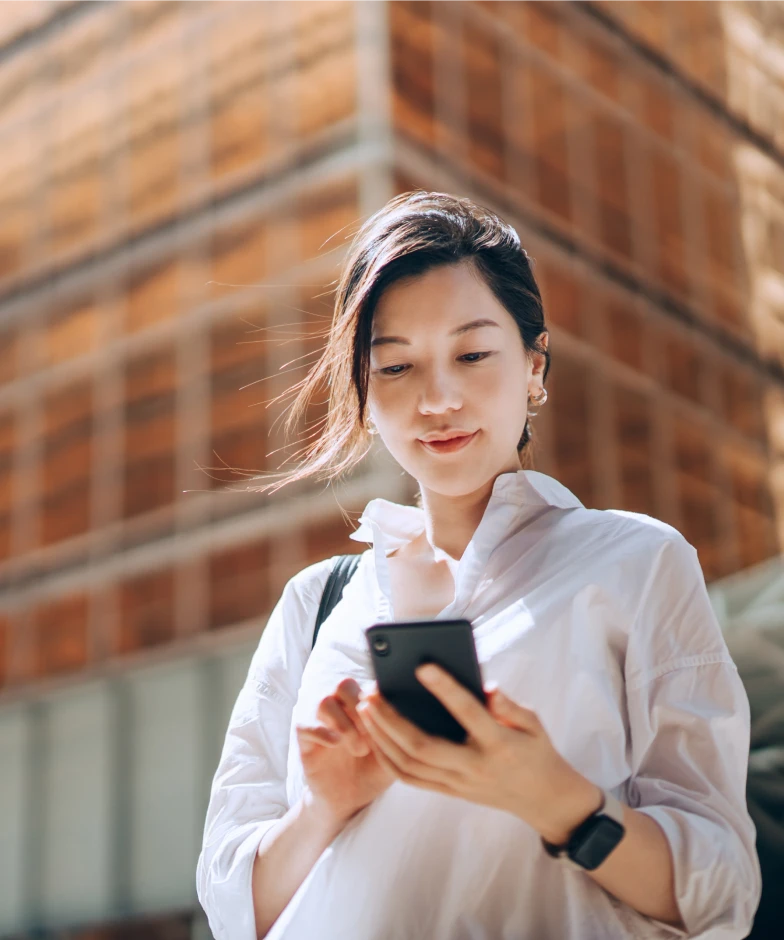 A woman looking at her mobile phone with an office building behind her. Login to Stifel Wealth Tracker.